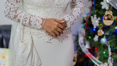 A person wearing an elegant white lace dress with intricate floral patterns, standing beside a decorated Christmas tree adorned with colorful lights and ornaments. The person's hands are adorned with rings and a bracelet.