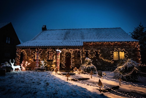 A cozy, snow-covered house is adorned with festive Christmas lights and decorations, including glowing reindeer and snowman figures. The lights illuminate the pathway and plants, creating a warm and inviting atmosphere amidst the evening sky.