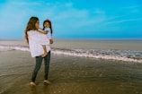 A person stands in shallow ocean water holding a young child. The sky is clear and blue, and gentle waves are rolling onto the shore. The person wears a white shirt and jeans, while the child is dressed in a light blue top with a pink crown.