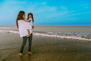 A person stands in shallow ocean water holding a young child. The sky is clear and blue, and gentle waves are rolling onto the shore. The person wears a white shirt and jeans, while the child is dressed in a light blue top with a pink crown.