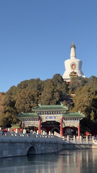 A traditional Chinese stupa stands atop a densely forested hill, overlooking an ornate, colorful gateway with green-tiled roofs. A stone bridge arches over a calm body of water in the foreground, with people walking across it.
