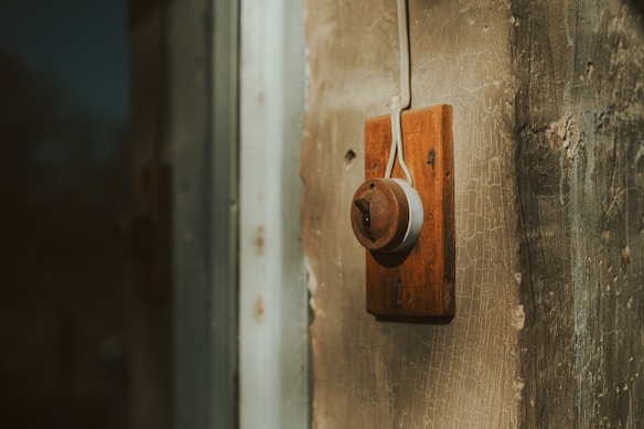 A vintage-style light switch mounted on a textured, aged wall. The switch has a wooden base with a rounded, ceramic top, featuring a single toggle. The wall displays visible cracks and an earthy, rugged texture.