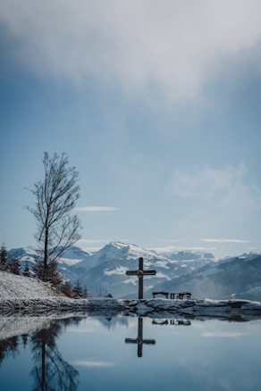 A serene mountain landscape with snow-covered peaks in the background. A cross is prominently placed in the foreground beside a still body of water, reflecting the sky and surrounding scenery. A solitary tree stands nearby, adding to the tranquility of the scene.