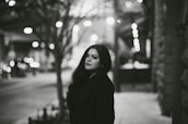 A woman stands on a dimly lit urban street, with blurred lights creating a bokeh effect in the background. She is looking directly at the camera with a calm expression. The setting appears to be night, and the photograph is in black and white.