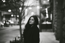 A woman stands on a dimly lit urban street, with blurred lights creating a bokeh effect in the background. She is looking directly at the camera with a calm expression. The setting appears to be night, and the photograph is in black and white.