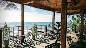 Open terrace overlooking a serene coastline with several ships in the distant sea. The area features wooden tables and chairs arranged neatly, potted plants, and a thatched roof offering a shaded seating area. The setting appears calm, airy, and relaxing, perfect for enjoying the scenic ocean view.
