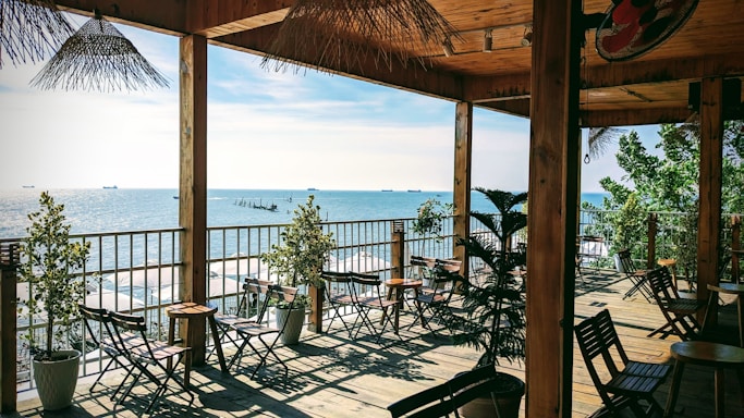 Open terrace overlooking a serene coastline with several ships in the distant sea. The area features wooden tables and chairs arranged neatly, potted plants, and a thatched roof offering a shaded seating area. The setting appears calm, airy, and relaxing, perfect for enjoying the scenic ocean view.