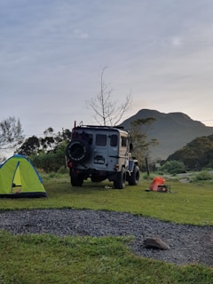 A rugged off-road vehicle is parked on a grassy campsite near a vibrant green and blue tent. In the background, there is a mountain and a leafless tree against a slightly overcast sky. On the ground, a few camping items, including an orange bag and a small black box, can be seen.
