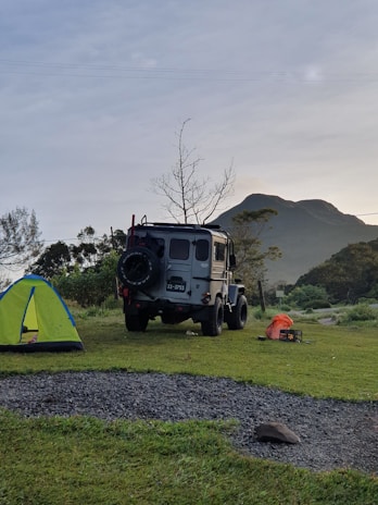 A rugged off-road vehicle is parked on a grassy campsite near a vibrant green and blue tent. In the background, there is a mountain and a leafless tree against a slightly overcast sky. On the ground, a few camping items, including an orange bag and a small black box, can be seen.