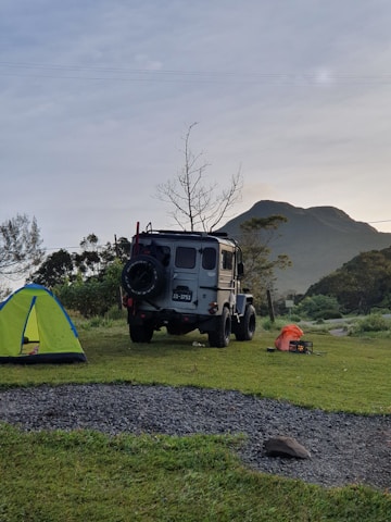 A rugged off-road vehicle is parked on a grassy campsite near a vibrant green and blue tent. In the background, there is a mountain and a leafless tree against a slightly overcast sky. On the ground, a few camping items, including an orange bag and a small black box, can be seen.