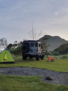 A rugged off-road vehicle is parked on a grassy campsite near a vibrant green and blue tent. In the background, there is a mountain and a leafless tree against a slightly overcast sky. On the ground, a few camping items, including an orange bag and a small black box, can be seen.