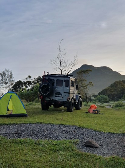 A rugged off-road vehicle is parked on a grassy campsite near a vibrant green and blue tent. In the background, there is a mountain and a leafless tree against a slightly overcast sky. On the ground, a few camping items, including an orange bag and a small black box, can be seen.