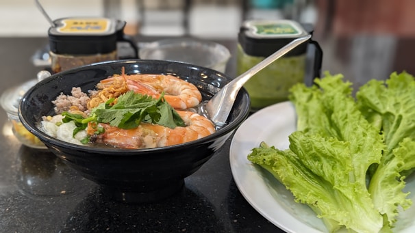 A black bowl filled with a flavorful soup featuring large shrimp, ground meat, garnished with fresh herbs. Beside it, a white plate holds crisp green lettuce leaves. Behind the bowl, jars contain condiments, possibly chili sauce and another sauce. A spoon rests inside the bowl ready for use.