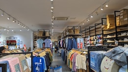 A retail clothing store interior with rows of neatly arranged shirts hanging on racks. Shelves above hold stacks of shoe boxes, and a variety of shoes are displayed on the right side. The store is well-lit with ceiling spotlights and has a clean, organized appearance.