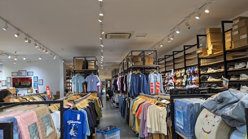A retail clothing store interior with rows of neatly arranged shirts hanging on racks. Shelves above hold stacks of shoe boxes, and a variety of shoes are displayed on the right side. The store is well-lit with ceiling spotlights and has a clean, organized appearance.