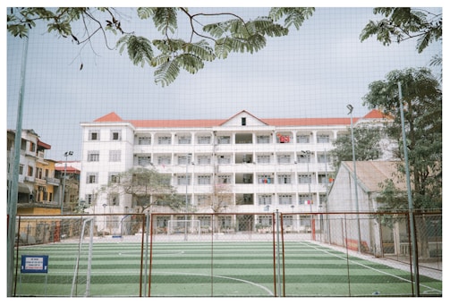 A large four-story building with a white facade and red roof stands behind a fenced sports field. The field has a green synthetic surface with white markings, likely for soccer. Trees partially frame the image, and a net is visible in the foreground.