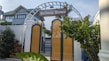 A decorative archway with a wooden sign reading 'Wellness Homes' stands above yellow gates. Lush greenery surrounds the entrance, with a modern house visible in the background under a clear sky.