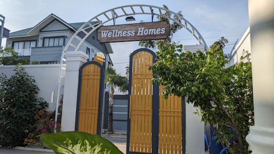 A decorative archway with a wooden sign reading 'Wellness Homes' stands above yellow gates. Lush greenery surrounds the entrance, with a modern house visible in the background under a clear sky.