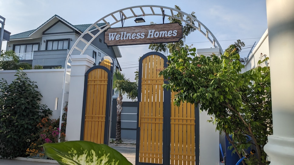 A decorative archway with a wooden sign reading 'Wellness Homes' stands above yellow gates. Lush greenery surrounds the entrance, with a modern house visible in the background under a clear sky.