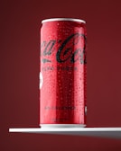 A red and black soda can, with visible condensation and droplets, sits upright on a white shelf against a deep red background. The can is labeled with the brand name and indicates it contains no sugar and no calories.