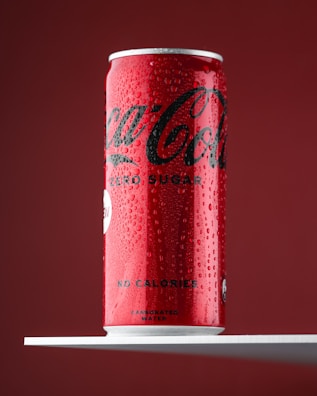 A red and black soda can, with visible condensation and droplets, sits upright on a white shelf against a deep red background. The can is labeled with the brand name and indicates it contains no sugar and no calories.