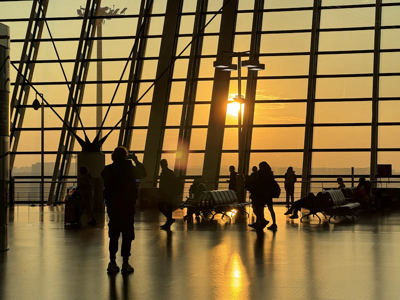 Travellers silhouetted against a golden sunrise through the tall windows of an airport terminal