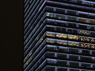 A modern high-rise building with glass windows lit in various colors, creating a grid pattern. The lights inside the building are in shades of blue and yellow, with some windows appearing dark. The structure's lines emphasize a sense of symmetry and order.