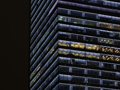 A modern high-rise building with glass windows lit in various colors, creating a grid pattern. The lights inside the building are in shades of blue and yellow, with some windows appearing dark. The structure's lines emphasize a sense of symmetry and order.