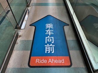 A directional sign on the floor with a blue arrow pointing forward and containing Chinese characters in white, and the English text 'Ride Ahead' in an orange section at the bottom. The sign is situated between glass walls, possibly in a transit station or walkway.