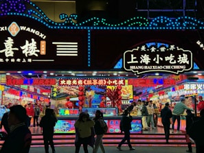 A vibrant and colorful street food market stall, illuminated with bright neon signs and lights. People are gathered around, some in conversation and others ordering food. The signs display Chinese characters, advertising a variety of snacks and delicacies.