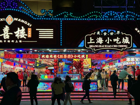 A vibrant and colorful street food market stall, illuminated with bright neon signs and lights. People are gathered around, some in conversation and others ordering food. The signs display Chinese characters, advertising a variety of snacks and delicacies.