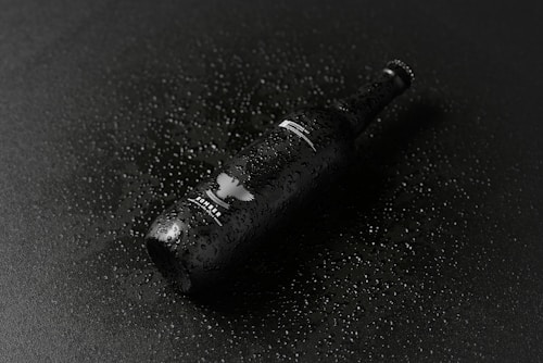 A dark glass beer bottle lying on a black surface covered with small droplets of water, creating a glossy and sleek appearance. The bottle displays a white logo and some text, enhancing the overall contrast with the dark tones.