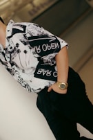 A person wearing a patterned shirt with black and white graphics stands casually, with one hand in their pocket and a gold watch visible on their wrist. The background is blurred, emphasizing the focus on the clothing and accessories.
