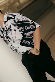 A person wearing a patterned shirt with black and white graphics stands casually, with one hand in their pocket and a gold watch visible on their wrist. The background is blurred, emphasizing the focus on the clothing and accessories.