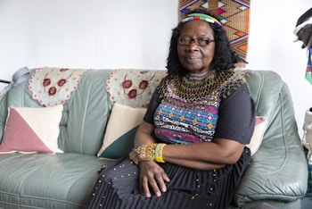 An elderly woman sits on a green leather couch adorned with embroidered doilies. She is wearing traditional colorful beaded jewelry and a dress with intricate patterns. The background includes patterned tapestry on the wall and a few pillows in pink and cream tones on the couch.