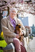 An elderly person wearing a tan coat and a purple shirt smiles while sitting on a stone ledge. In their arms, they hold a small brown dog. Blooming cherry blossom trees with pink flowers and a clear sky are in the background, suggesting springtime. The setting appears to be a peaceful suburban street lined with greenery.