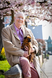 An elderly person wearing a tan coat and a purple shirt smiles while sitting on a stone ledge. In their arms, they hold a small brown dog. Blooming cherry blossom trees with pink flowers and a clear sky are in the background, suggesting springtime. The setting appears to be a peaceful suburban street lined with greenery.