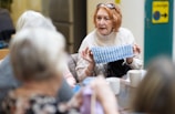 An older woman is sitting at a table in a communal setting, displaying a piece of knitted fabric to others. She is wearing a cream-colored sweater and glasses perched on her head. Around the table, there are several other people engaged in the activity, with cups and yarn visible on the table.