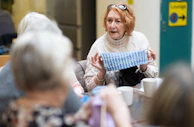 Smiling community members sharing their crochet creations during a casual meet-up in a warm, wood-paneled room.