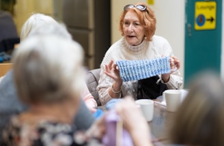 An older woman is sitting at a table in a communal setting, displaying a piece of knitted fabric to others. She is wearing a cream-colored sweater and glasses perched on her head. Around the table, there are several other people engaged in the activity, with cups and yarn visible on the table.