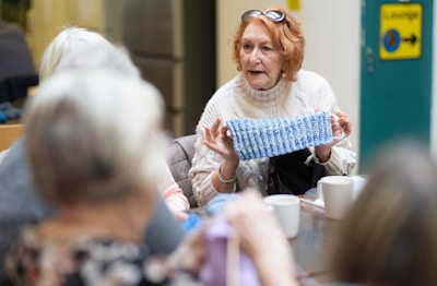 An older woman is sitting at a table in a communal setting, displaying a piece of knitted fabric to others. She is wearing a cream-colored sweater and glasses perched on her head. Around the table, there are several other people engaged in the activity, with cups and yarn visible on the table.