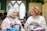 Two elderly women are sitting together in a cozy indoor setting, engaged in knitting. They appear to be in conversation, one with short white hair wearing a scarf and the other with short red hair in a sweater. Both have knitting needles and yarn in front of them. The background features soft-focus people and arched windows, suggesting a warm, community environment.