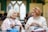 Two elderly women are sitting together in a cozy indoor setting, engaged in knitting. They appear to be in conversation, one with short white hair wearing a scarf and the other with short red hair in a sweater. Both have knitting needles and yarn in front of them. The background features soft-focus people and arched windows, suggesting a warm, community environment.