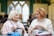 Two elderly women are sitting together in a cozy indoor setting, engaged in knitting. They appear to be in conversation, one with short white hair wearing a scarf and the other with short red hair in a sweater. Both have knitting needles and yarn in front of them. The background features soft-focus people and arched windows, suggesting a warm, community environment.
