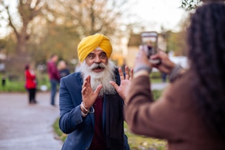 A bearded man wearing a yellow turban and a blue suit appears to be animatedly talking or gesturing, possibly while another person takes a photo of him using a smartphone. The background shows a park-like setting with trees and other blurred figures.