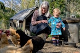 A person with long gray hair, wearing glasses and a dark apron, is crouching next to a small child dressed in a colorful outfit with rainbow-striped leggings. They are surrounded by several chickens in an outdoor setting, with a chicken coop and some metal containers in the background. There's a sense of warmth and care as the adult gently holds the child’s hand.