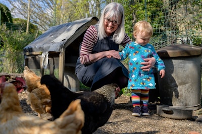 A person with long gray hair, wearing glasses and a dark apron, is crouching next to a small child dressed in a colorful outfit with rainbow-striped leggings. They are surrounded by several chickens in an outdoor setting, with a chicken coop and some metal containers in the background. There's a sense of warmth and care as the adult gently holds the child’s hand.