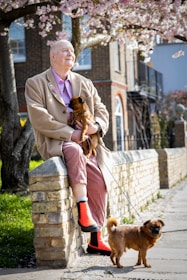 An elderly person sits on a brick wall under blooming cherry blossom trees. They are holding a small brown dog and a second similar dog is on a leash, standing beside them. The person is wearing a beige coat, pink shirt, and bright orange boots. The background includes a path and a residential building.