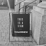 A black signboard is positioned on a brick pavement near a stone staircase. The board reads 'THIS IS A SIGN' with 'YOGARENEW' written below. The surrounding area includes architectural stone details and a metal railing.