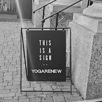 A black signboard is positioned on a brick pavement near a stone staircase. The board reads 'THIS IS A SIGN' with 'YOGARENEW' written below. The surrounding area includes architectural stone details and a metal railing.